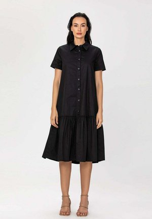 Femme portant une robe noire boutonnée jusqu'aux genoux avec des manches courtes et un ourlet volanté, debout devant un fond blanc uni.