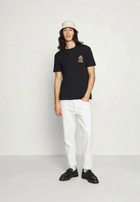 T-shirt preta com um pequeno logotipo bege no lado esquerdo do peito, combinada com calças brancas e sapatos pretos, usada por um modelo de cabelo comprido.