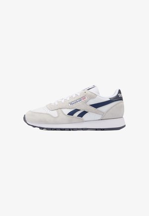 Witte en lichtgrijze Reebok sneaker met marineblauwe strepen, suède details en witte veters op een witte achtergrond.
