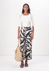 Cardigan en maille blanche légère sur un haut blanc ajusté, associé à un pantalon large rayé de zèbre noir et crème. Un sac tressé complète le look.