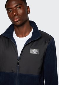 Veste en polaire marine avec fermeture éclair, présentant un panneau en tissu noir texturé, un col montant et une patch logo sur le côté supérieur gauche.
