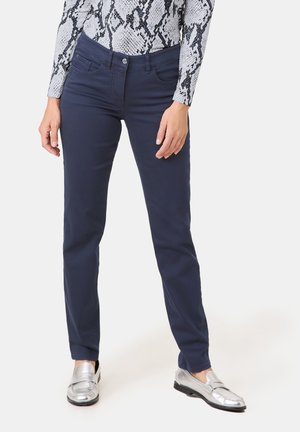 Femme portant un pantalon droit bleu marine, des mocassins argentés et un haut à manches longues imprimé serpent, debout les mains détendues le long du corps.