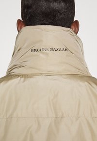 Beige puffervest med høj krave, prydet med teksten "BRUUNS BAZAAR" på ryggen i sort. Glat, blank tekstur og minimalistisk design.