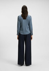 Blauwe zijden blouse met lange mouwen, voorzien van sleuven bij de manchetten, gecombineerd met wijde marineblauwe broek, weergegeven vanuit de achterkant.