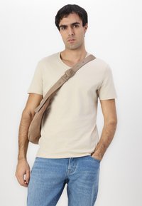 Beige Baumwoll-T-Shirt mit V-Ausschnitt, kombiniert mit blauen Jeans und einer braunen Umhängetasche mit einem verstellbaren Riemen.