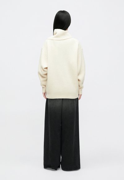 Prevelik kremast pleteni pulover z visokim ovratnikom, v kombinaciji s širokimi črnimi hlačami. Pulover z rebrasto teksturo, hlače iz gladkega materiala.