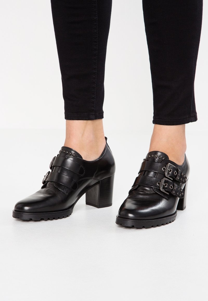 Pieds portant des chaussures à talons en cuir noir avec doubles brides à boucles et détails cloutés, associées à un pantalon noir ajusté.