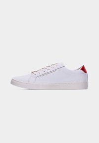 Baskets basses blanches avec lacets blancs, languette rouge au talon et logo doré "Tommy Hilfiger" sur le côté sur un fond uni.