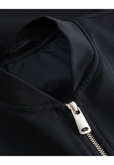 Gros plan d'une veste en tissu noir avec une fermeture éclair argentée partiellement ouverte près du col.