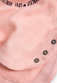 Tela rosa con una textura suave que presenta botones metálicos decorativos a lo largo del borde, con costuras en contraste y una etiqueta impresas en la parte superior.
