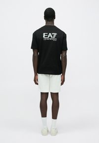 Férfi áll háttal, fekete EA7 Emporio Armani pólót, fehér rövidnadrágot, fehér zoknit és fehér tornacipőt viselve, egyszínű háttér előtt.