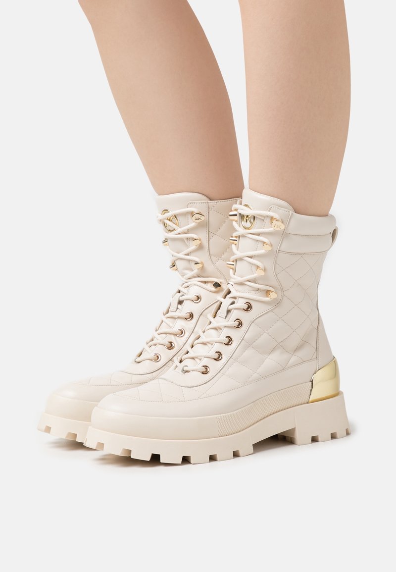Bottes chevilles en cuir crème matelassé à lacets, avec semelles épaisses, œillets dorés et accents métalliques dorés sur le talon, portées sur des jambes nues.