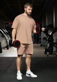 Un homme soulevant des haltères noirs avec des accents rouges, portant un t-shirt et un short de sport beige, associés à des baskets blanches sur le sol d'une salle de sport.