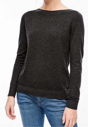 Frau trägt einen eng anliegenden dunkelgrauen Langarmpullover und blaue Jeans, dargestellt von Hals bis Hüfte vor weißem Hintergrund.