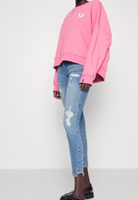 Różowy oversize'owy sweatshirt z białym logo, w komplecie z przecieranymi jasnoniebieskimi dżinsami i czarnymi butami na platformie, wyeksponowany z boku.