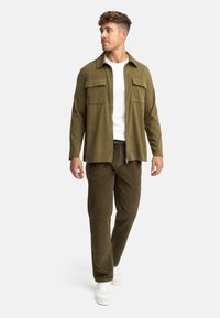 Chaqueta de pana verde oliva con dos bolsillos en el pecho, usada sobre una camiseta blanca; combinada con pantalones texturizados a juego y zapatillas blancas.
