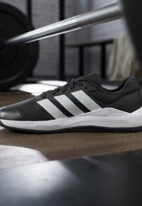 Chaussure de course Adidas noir et blanc sur le sol de la salle de sport avec une barre d’haltérophilie en arrière-plan flou, suggérant un cadre d’entraînement.