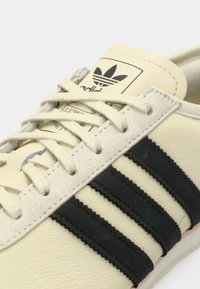 Αθλητικό παπούτσι από κρέμ δέρμα με μαύρες ρίγες, υφή και μπεζ κορδόνια. Διαθέτει ετικέτα στη γλώσσα με το λογότυπο σε μαύρο και λευκό.