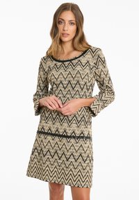 Beiges Kleid mit einem schwarzen Zickzack-Muster, rundem Ausschnitt und Dreiviertelärmeln. Strukturierter Stoff mit lockerer Passform.