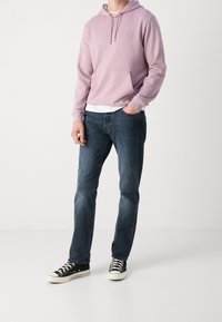 Jasnofioletowa bluza z kapturem z kieszenią kangurką, nałożona na białą koszulę. Do tego ciemnoniebieskie dżinsy i czarne trampki z białą podeszwą.