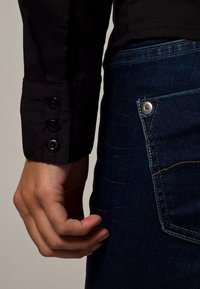 Camicia nera con bottoni ai polsini, abbinata a jeans denim blu scuro con una tasca visibile e dettaglio in metallo sul bottone.