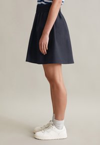 Donkerblauwe A-lijn rok met een elastische tailleband, zijwaarts weergegeven, gecombineerd met witte sneakers en grijze sokken. Glad van textuur.