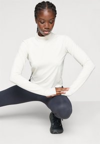 Vrouw in een witte lange mouwtop en zwarte leggings die haar benen strekt, naar beneden kijkend met haar handen gekruist over één knie.