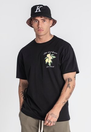 La camiseta de algodón negra presenta un gráfico frontal de limones amarillos con hojas verdes. El modelo lleva un sombrero de pescador negro con un logo blanco en forma de "K".