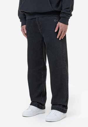 Personne portant un jean noir à jambes larges avec un petit logo brodé et des baskets blanches, debout devant un fond uni.