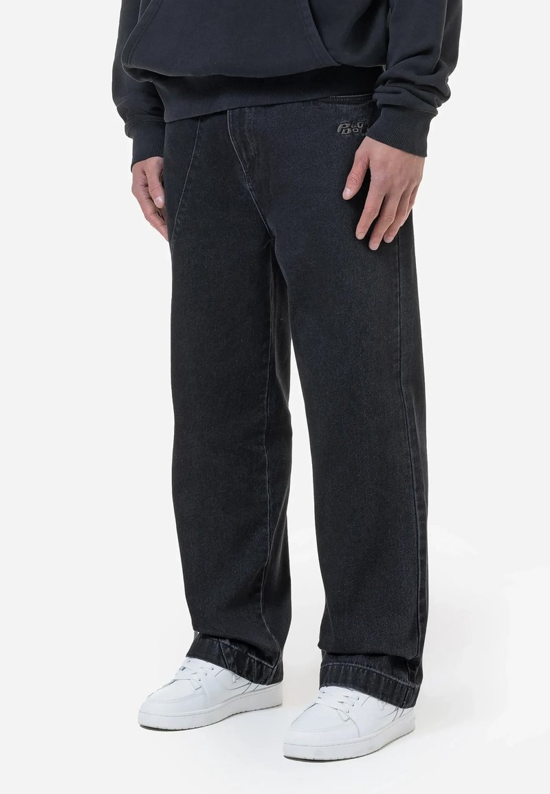 Personne portant un jean noir à jambes larges avec un petit logo brodé et des baskets blanches, debout devant un fond uni.