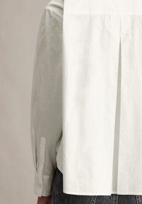 Witte button-up shirt met een geplooide achterkant en lange mouwen. Het materiaal lijkt lichtgewicht met een gladde textuur. De manchetten hebben knoopsluitingen.