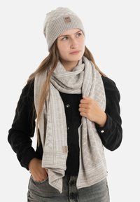 Gebreide grijze beanie met een gestructureerd patroon en een logopatch, gecombineerd met een bijpassende gestructureerde sjaal, gedragen over een zwart top.