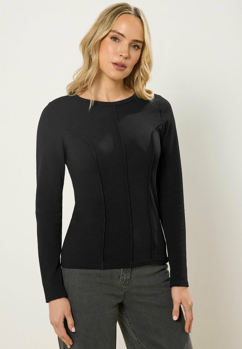 Femme aux cheveux blonds ondulés, portant un haut noir à manches longues ajusté et un pantalon gris, debout devant un fond clair uni.