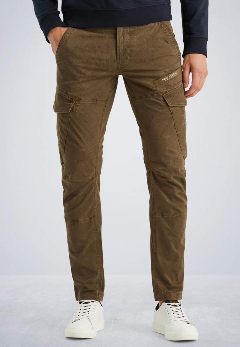 Bruine cargobroek van textielstof, met meerdere zakken en zichtbare stiksels. Draagt met witte sneakers voor contrast.