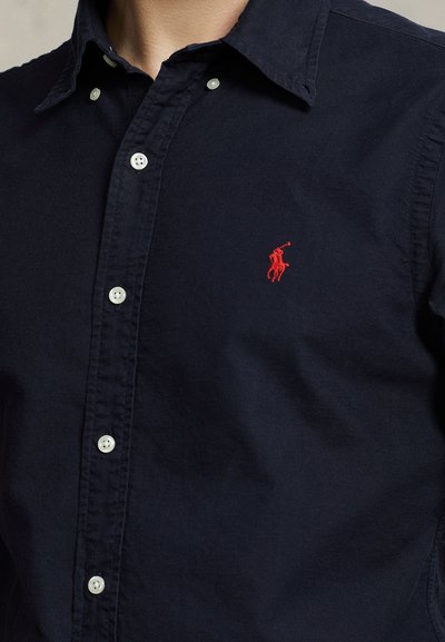 Primo piano di una camicia blu scuro con bottoni bianchi e un piccolo logo ricamato rosso di un giocatore di polo sul petto.