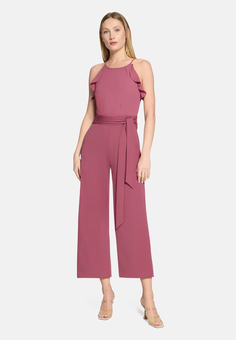 Rosa ärmelloser Jumpsuit mit Rüschen-Trägern, tailliertem Gürtel und weitem Bein aus glattem Stoff. Klare Sandalen mit Absatz vervollständigen den Look.