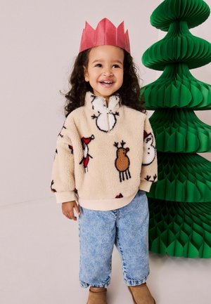 Fleece-Pullover in Beige mit festlichen Tiermotiven, hohem Kragen und Reißverschluss. Kombiniert mit blauen Jeans und braunen Stiefeln, ergänzt durch eine rote Papiertüte.