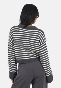Vrouw met lang zwart haar, die een grijs-witte horizontaal gestreepte trui en donkere grijze broek draagt, met haar rug naar de camera gekeerd op een effen achtergrond.