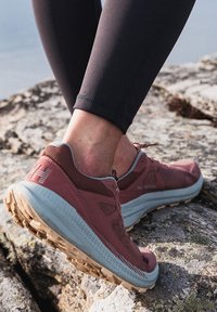 Zapatilla de running ligera en rojo-rosado con parte superior de malla, acentos grises y suela de goma texturizada. Ideal para actividades al aire libre, mostrada sobre una superficie rocosa.