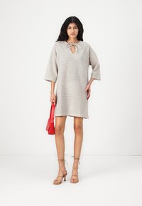 Vestido bege xadrez com decote em gota, mangas três quartos e um corte solto, combinado com uma pequena bolsa vermelha.