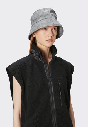 Chapeau seau gris avec une texture lisse et des détails cousus. Porté avec un gilet en polaire noir avec une fermeture éclair à l'avant et un col haut.