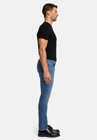 Schwarzes T-Shirt mit kurzen Ärmeln, hellblau Slim-Fit-Jeans und schwarze Schnürschuhe; Person im Profil stehend.