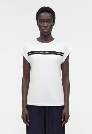 Γυναίκα που στέκεται μπροστά φοράει λευκό αμάνικο τοπ με μαύρη οριζόντια λωρίδα λογότυπου και άνετο σκούρο παντελόνι.