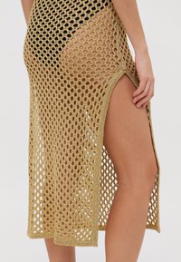 Zelta, vaļīga adīta krošu pārklājums ar sānu šķēlumu, kas piedāvā brīvu, gaisu caurlaidīgu tekstūru un vidēja garuma dizainu, kas smuki krīt līdz gurniem.