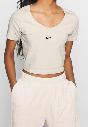Woman wearing a light beige cropped Nike t-shirt with short sleeves and high-waisted white pants, hands in pockets.