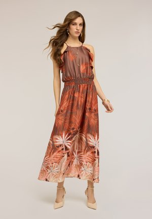 Femme aux longs cheveux bruns portant une robe maxi sans manches à motifs tropicaux marron et orange, et des talons beiges, se tenant debout avec la main légèrement levée.