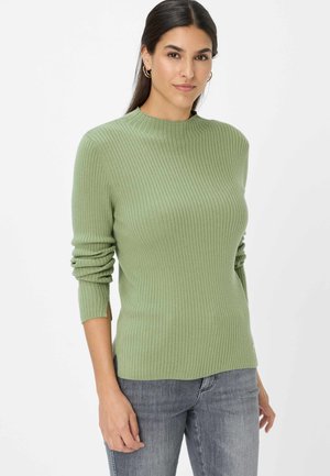 Grüner, gerippter Pullover mit hohem Kragen und langen Ärmeln, ausgestattet mit umgeschlagenen Manschetten und seitlichen Schlitzen. Kombiniert mit grauen Jeans.