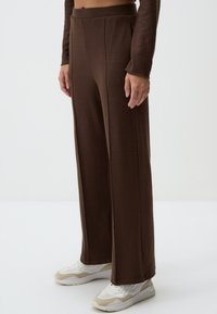 Braune, weite Hose aus einem strukturierten Stoff, mit hoher Taille und seitlichen Schlitzen, kombiniert mit weißen und beigen Sneakern.