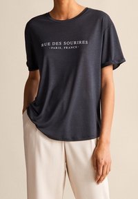 Dunkelgraues T-Shirt mit kurzen Ärmeln und lockerer Passform, mit weißem Text "RUE DES SOURIRES - PARIS, FRANCE" zentriert auf der Vorderseite.