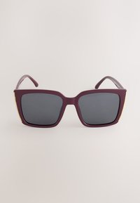 Neliönmuotoiset aurinkolasit, joissa paksut burgundinväriset kehykset ja tummennetut linssit yksivärisellä taustalla.